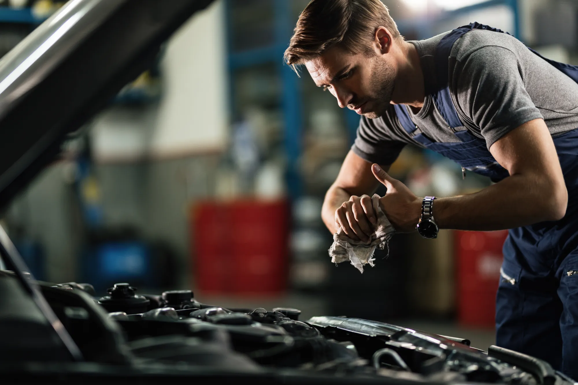 A mechanic in navy overalls wipes his hands with a rag while leaning over the open hood of a car in a repair shop.