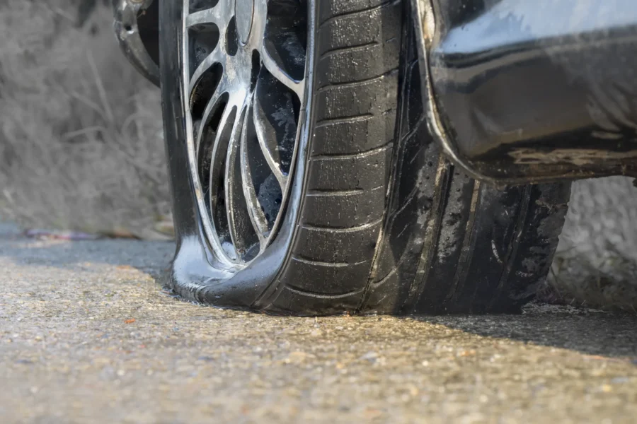 Close-up of a flat, deflated tire on an alloy wheel of a dark car resting on wet asphalt.